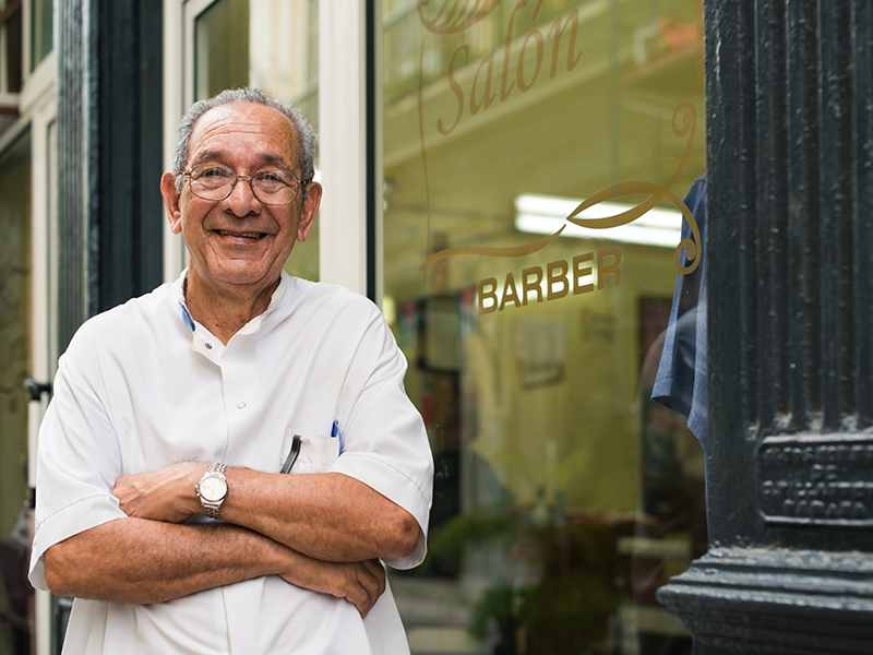 senior hispanic barber in old fashion barber's shop, posing and looking at camera with arms crossed near shop window