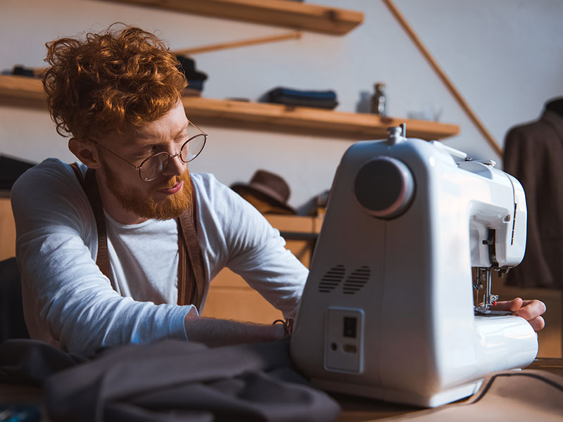 Focused young fashion designer in eyeglasses working with sewing machine