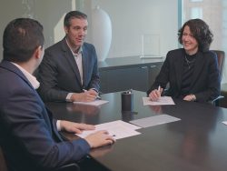 : Two businessmen and a businesswoman sit at a conference table.