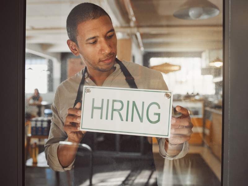 Business owner looking to hire new staff. Young businessman hanging a hiring sign in his shop entrance. Boss working in coffeeshop hiring new employees. Hr employee hanging hiring signage in shop